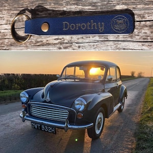 May include: A blue leather keychain with the name "Dorothy" engraved on it. The keychain is attached to a silver key ring. The keychain is sitting on a wooden surface. In the background, a classic blue car is parked on a road with a sunset in the background.