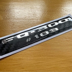 Peut inclure: Un autocollant noir et argent avec le texte "F-150 #183" sur un fond en fibre de carbone.