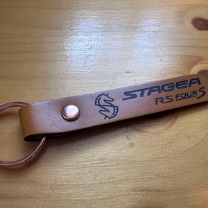 Op de afbeelding: Een bruine leren sleutelhanger met een metalen ring. De sleutelhanger heeft de tekst "STAGEA RS FOUR S" en een gestileerd paardenlogo dat erop is gegraveerd.