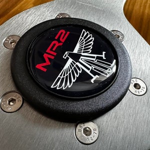 May include: Close-up of a car steering wheel centre cap. The cap is black with the red text "MR2" and a white graphic of a bird. The cap is surrounded by a brushed metal plate and secured with silver bolts.