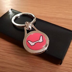 Op de afbeelding: Een zilveren sleutelhanger met een roze cirkel met een wit logo. De sleutelhanger ligt op een zwarte achtergrond.