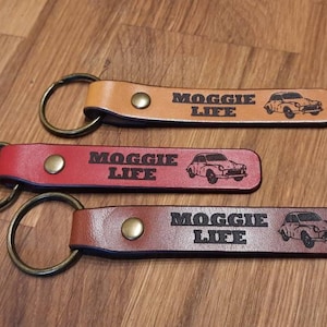 May include: Three leather keychains with a small car design and the text "Moggie Life" in black lettering. The keychains are in brown, red, and tan.