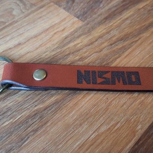 Op de afbeelding: Bruine leren sleutelhanger met een zilveren sleutelring en het woord "NISMO" in zwart gegraveerd.