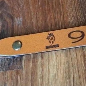 Könnte beinhalten: Ein brauner Leder-Schlüsselanhänger mit einem Metallring. Der Schlüsselanhänger hat ein Saab-Logo und die Nummer 95 eingraviert.