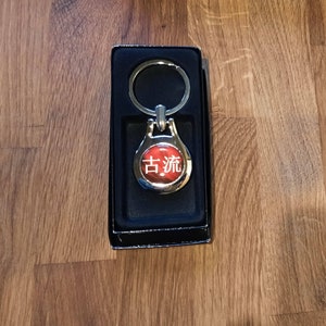 Pode incluir: Um chaveiro de prata com um charme circular vermelho com os caracteres chineses "古流" em branco.