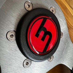 May include: A close-up of a black steering wheel hub with a red and black Mazda logo. The hub is made of metal and has six silver screws.