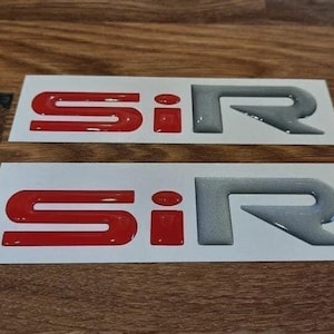 May include: Two SiR emblem stickers. The "Si" is red, and the "R" is silver. The stickers are on a white background, and the background is a wooden surface.
