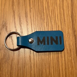 Könnte beinhalten: Blauer Leder-Schlüsselanhänger mit einem silbernen Ring und dem Wort "MINI" in schwarzen Buchstaben.
