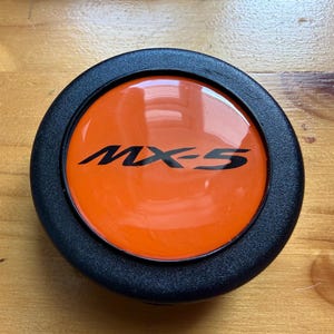 Peut inclure: Un bouton de klaxon circulaire avec un anneau extérieur noir texturé et un insert central orange brillant. Le texte MX-5 est imprimé en noir au milieu de la surface orange. L'objet présente une finition lisse et réfléchissante sur la partie centrale.