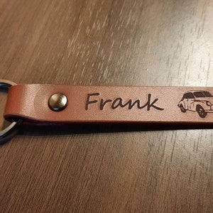 May include: Brown leather keychain with a silver ring and a small car design engraved on it. The keychain is personalized with the name "Frank".