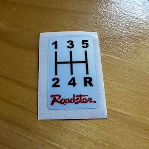 Könnte beinhalten: Weißer Aufkleber mit einem schwarzen Schaltmuster und dem Wort "Roadster" in Rot. Das Schaltmuster zeigt die Zahlen 1, 2, 3, 4, 5 und den Buchstaben R.