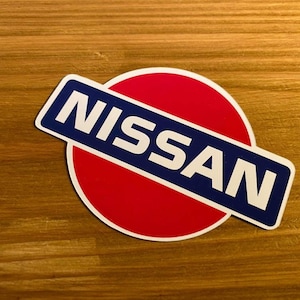 Può includere: Un logo rosso e blu con la parola "NISSAN" in lettere bianche.