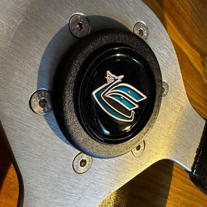 Könnte beinhalten: Ein silbernes Lenkrad mit einer schwarzen Mittelnabe, die ein blaues und weißes Logo trägt. Die Nabe ist mit sechs silbernen Schrauben befestigt.