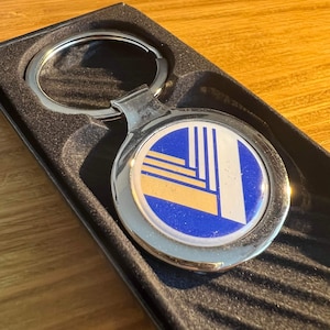 Peut inclure: Un porte-clés argenté avec un emblème rond orné d'un motif géométrique bleu, blanc et doré. Le porte-clés est présenté dans un coffret cadeau noir texturé, sur une surface en bois.