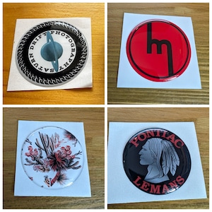 May include: Four round stickers with different designs. The first sticker is black and white with the text "Saturn Drift Photography". The second sticker is red with a black "M" in the centre. The third sticker is white with a red and green cactus design. The fourth sticker is black with red text that says "Pontiac Lemans".
