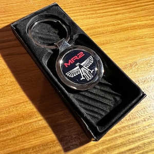 May include: A silver keychain with a black circular charm featuring a white eagle and the red text "MR2". The keychain is in a black box with a textured pattern.