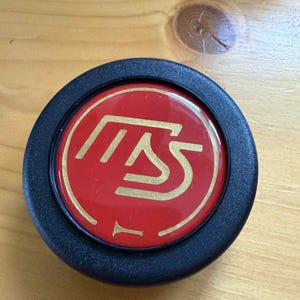 May include: A circular black object with a red central disc. The disc displays a gold-coloured graphic design. This is likely a button or emblem, possibly for a vehicle or piece of machinery.