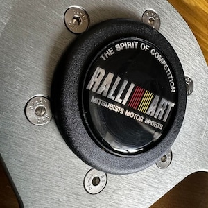 May include: Close-up of a silver steering wheel with a black centre cap. The cap has a logo that reads "The Spirit of Competition RALLIART Mitsubishi Motor Sports".