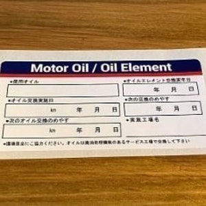 Op de afbeelding: Een witte sticker met blauwe en rode randen, met de tekst "Motor Oil / Oil Element". De sticker heeft velden om de data en kilometerstand van de olieverversing te registreren.