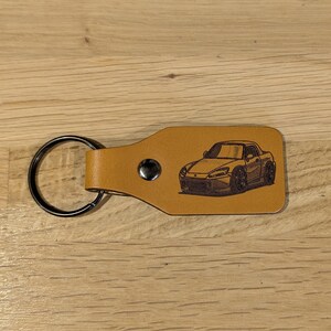 Può includere: Un portachiavi in ​​cuoio marrone con un anello portachiavi argentato e un'immagine incisa al laser di una Honda S2000 argentata.