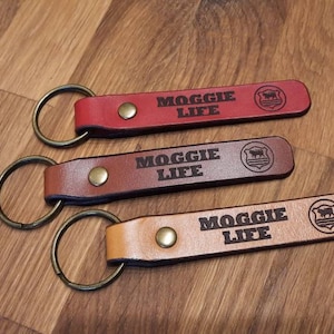 May include: Three leather keychains with a metal ring. Each keychain has the text "MOGGIE LIFE" and a small logo of a dog in a shield. The keychains are in red, brown, and tan.