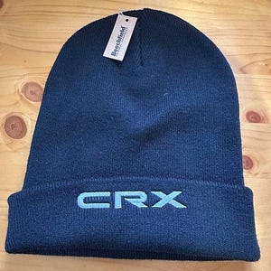May include: A navy blue beanie hat with the embroidered letters "CRX" in a lighter blue. The hat has a folded brim and a tag that reads "Beechfield". The beanie is made of a soft, knitted material.