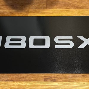 Könnte beinhalten: Ein schwarzes rechteckiges Schild mit dem Text "180SX" in silbernen Buchstaben.