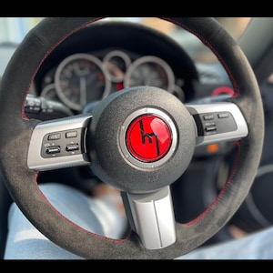 Può includere: Volante nero con cuciture rosse e un emblema rosso. Il volante presenta pulsanti di controllo argentati e un'impugnatura strutturata. Il cruscotto e gli indicatori dell'auto sono visibili sullo sfondo.