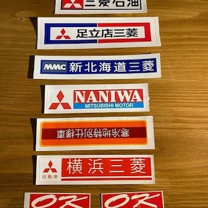 Peut inclure: Un ensemble de huit autocollants de concessionnaire automobile Mitsubishi d'époque. Les autocollants sont blancs avec du texte et des logos rouges, bleus et noirs. Les autocollants présentent le logo Mitsubishi et le texte "Mitsubishi", "Naniwa", "Mitsubishi Motor", "Ok", "New Hokkaido Mitsubishi", "Yokohama Mitsubishi", "Adachi Mitsubishi" et "Mitsubishi Oil".