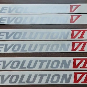 Puede incluir: Seis pegatinas rectangulares con la palabra "EVOLUTION" en gris y números romanos V, VI y VII en rojo. Las pegatinas están sobre una superficie marrón oscuro.