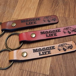 May include: Three leather keychains in brown, red, and tan. Each keychain has a metal ring and a rectangular leather tag with the text "MOGGIE LIFE" and a car graphic. The keychains are on a wooden surface.