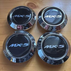 Peut inclure: Quatre enjoliveurs de roue circulaires chromés, avec le texte "MX-5" en blanc sur fond noir. Les enjoliveurs sont brillants et réfléchissants, disposés sur une surface en bois.