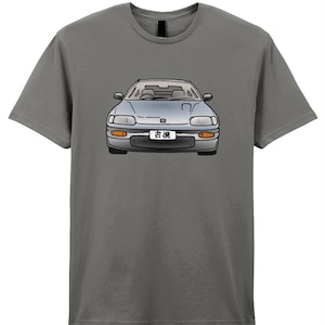May include: Gray t-shirt featuring a front-facing illustration of a silver car with orange headlights and a Japanese license plate. The car is centered on the shirt. The shirt is a solid gray color.