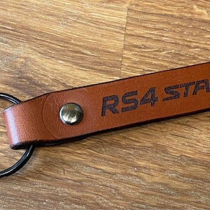 May include: Brown leather keychain with a black metal ring and the text "RS4 STAGEA" engraved in black.