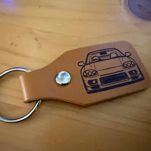 Pode incluir: Um porta-chaves de couro castanho claro com uma argola e um botão de pressão prateados. O porta-chaves tem um desenho a preto de um carro.