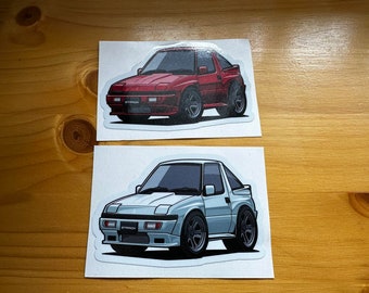 May include: Two stickers featuring cartoon illustrations of a red and a silver Mitsubishi Starion. The stickers are on a wooden surface.