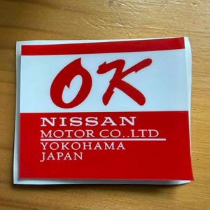 Può includere: Un adesivo rettangolare con un design bianco e rosso. La sezione superiore è bianca con le lettere rosse "OK". La sezione inferiore è rossa con il testo "NISSAN MOTOR CO. LTD YOKOHAMA JAPAN".