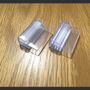 May include: Two transparent plastic sign holders are shown on a wooden surface. Each holder has a rectangular base and a curved top section designed to hold a sign. The holders are clear, allowing the wooden grain to be visible.