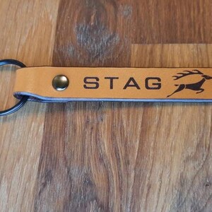 May include: A tan leather keychain with a black metal ring. The keychain has the word "STAG" in dark brown letters, along with a silhouette of a deer. A metal rivet secures the leather to the ring.
