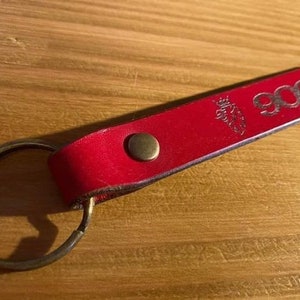Pode incluir: Um porta-chaves de couro vermelho com uma argola de metal. O porta-chaves tem o número "900" em relevo e um pequeno emblema. Um rebite cor de latão prende o couro à argola.