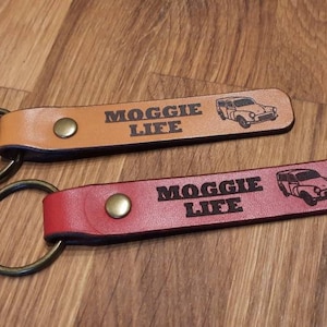May include: Two leather keychains with a metal ring. Both keychains have the text "MOGGIE LIFE" and a small image of a car. One keychain is brown and the other is red.