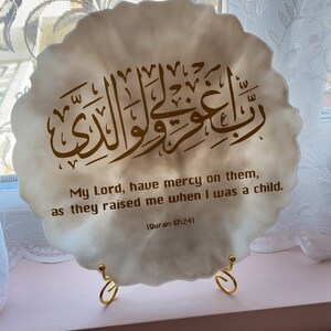 May include: A white, round, decorative plaque with a gold Arabic inscription and the English translation "My Lord, have mercy on them, as they raised me when I was a child. (Quran 17:24)". The plaque is on a gold metal stand.