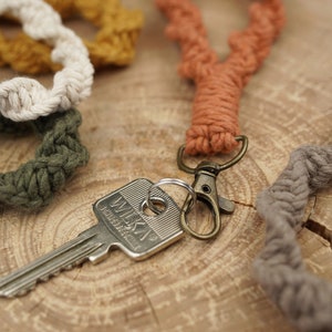 Könnte beinhalten: Ein Set aus vier handgefertigten Makramee-Schlüsselanhängern in verschiedenen Farben, darunter Beige, Grün, Orange und Braun. Die Schlüsselanhänger sind aus dickem, gedrehtem Kordel gefertigt und haben einen Schlüsselring aus Metall.
