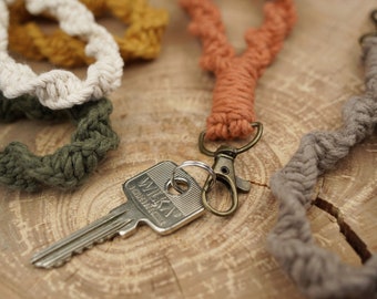 May include: A set of four handmade macrame keychains in various colours, including beige, green, orange, and brown. The keychains are made with thick, twisted cord and feature a metal key ring.