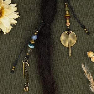 May include: A black hair braid with a beaded and charm hair tie. The tie features a round brass charm, a small spike charm, and a variety of colored beads.