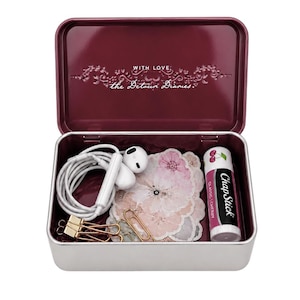 May include: A rectangular burgundy metal tin with the text "WITH LOVE, the Detour Diaries" printed on the inside lid. The tin contains white wired earbuds, a stack of floral-patterned paper cutouts, gold metal binder clips, and a cherry ChapStick.