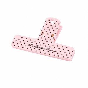 May include: A light pink plastic bag clip featuring a pattern of small dark brown polka dots. The text "the Detour Diaries" is printed in a dark brown cursive script across the center of the clip. It has a spring-loaded hinge mechanism.