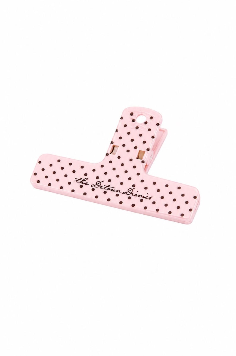 May include: A light pink plastic bag clip featuring a pattern of small dark brown polka dots. The text "the Detour Diaries" is printed in a dark brown cursive script across the center of the clip. It has a spring-loaded hinge mechanism.