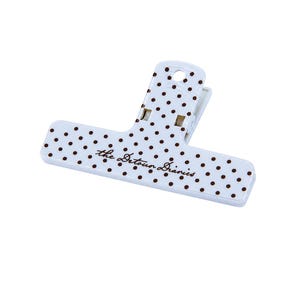 May include: A white plastic binder clip featuring a pattern of small brown polka dots across its surface. The text "the Detour Diaries" is printed in a brown cursive script on the front face of the clip, which includes a top handle hole.