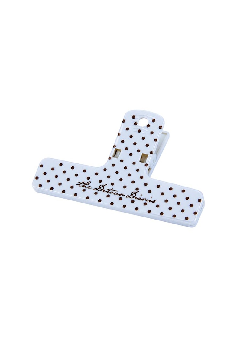 May include: A white plastic binder clip featuring a pattern of small brown polka dots across its surface. The text "the Detour Diaries" is printed in a brown cursive script on the front face of the clip, which includes a top handle hole.